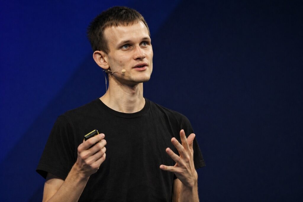 Ethereum co-founder Vitalik Buterin speaks onstage, holding a presentation clicker and gesturing expressively against a dark blue backdrop