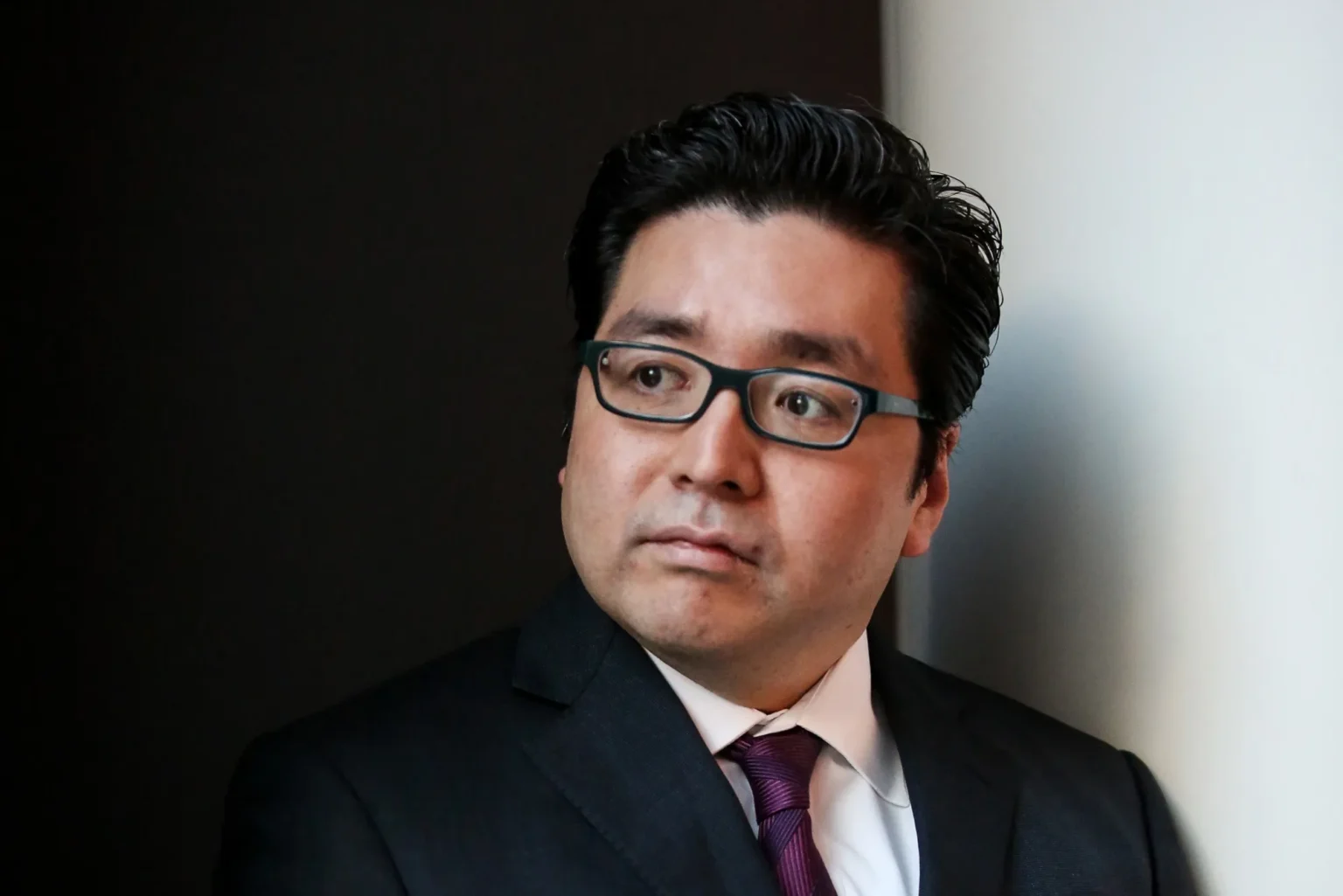 Tom Lee wearing glasses and a dark suit looks thoughtfully to the side, posed indoors against a contrasting black-and-white background