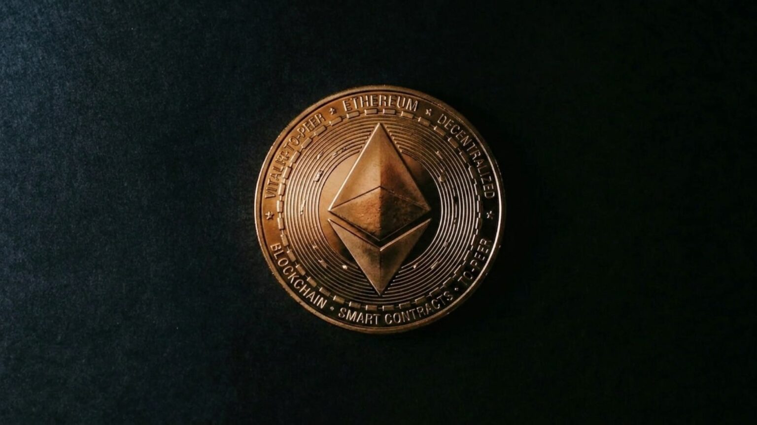 Close-up of a bronze Ethereum cryptocurrency coin on a dark textured surface, featuring a raised diamond logo and blockchain-themed lettering