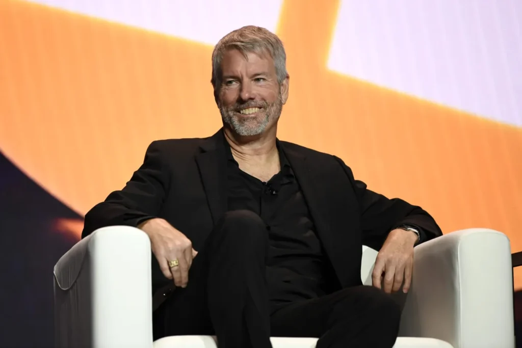 Bitcoin Has an Obituary Problem, and Michael Saylor Is Not Buying It Michael Saylor in a black suit sits in a white armchair onstage against bold orange and black graphics