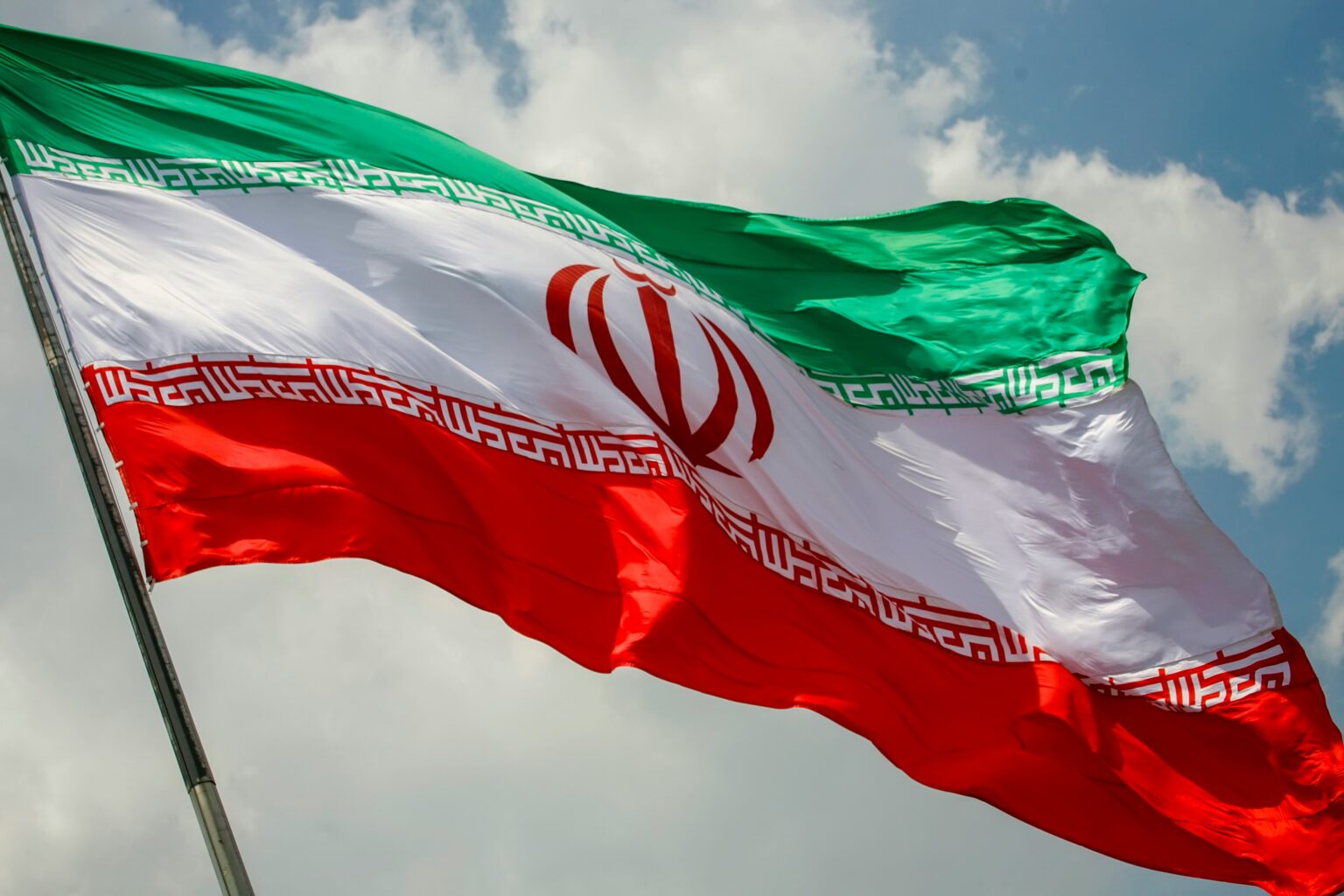 Iranian flag waving dramatically on a pole against a partly cloudy blue sky, its green, white, and red bands rippling in the wind