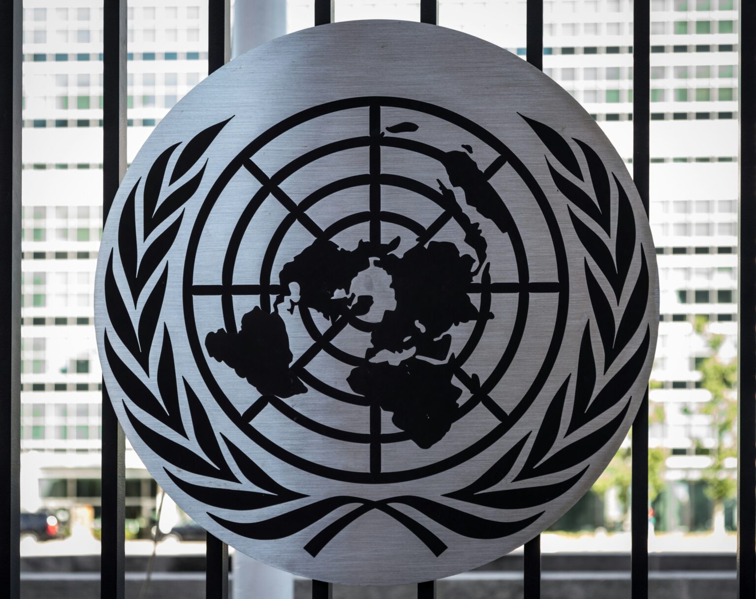 Bitcoin Gets a Seat at the UN’s Digital Development Table Close-up of the United Nations emblem: a world map encircled by latitude lines and olive branches, silhouetted against city windows