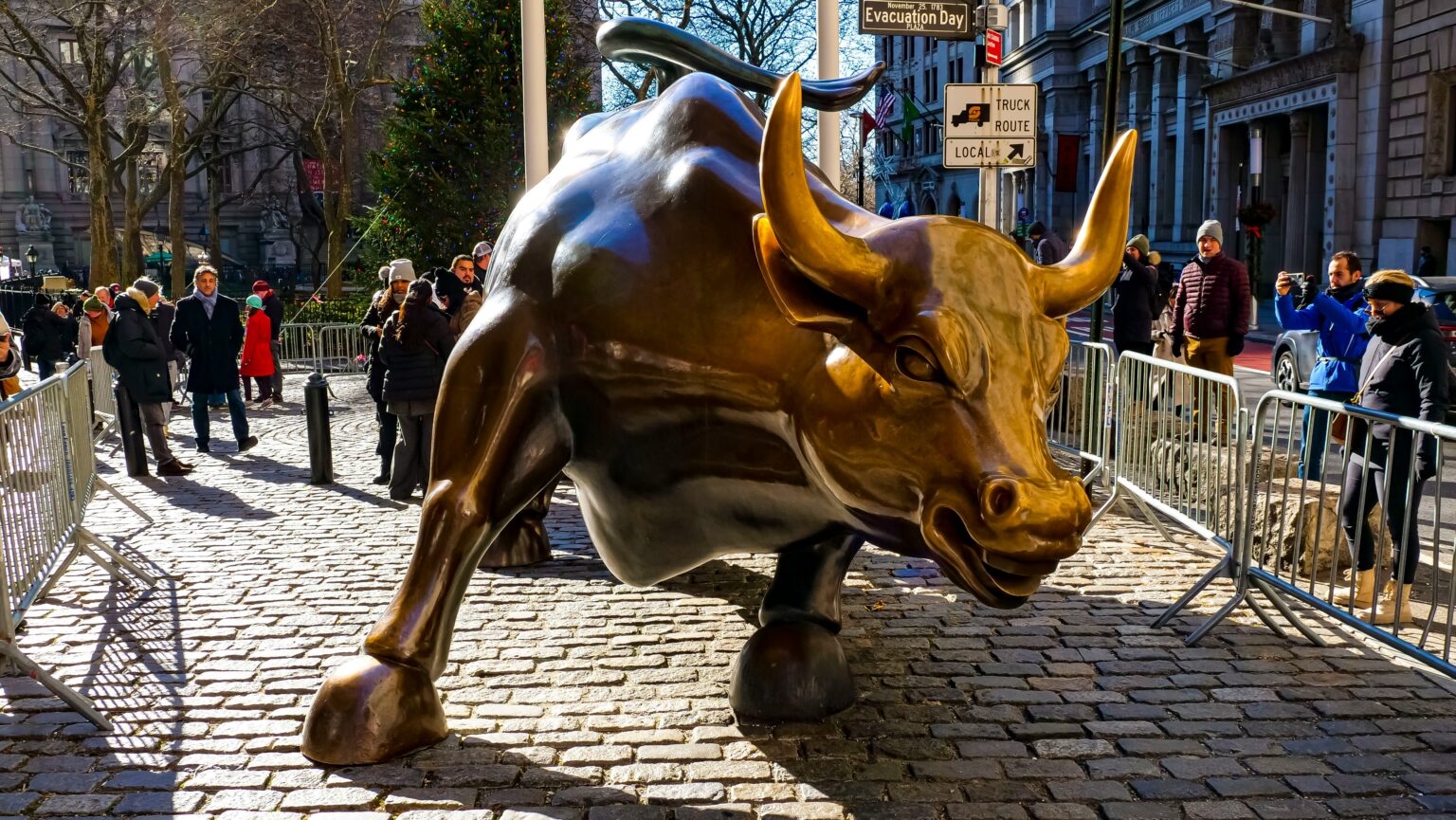 Charging bronze bull sculpture dominates a sunlit cobblestone street on Wall Street, NYC, as tourists gather behind barricades