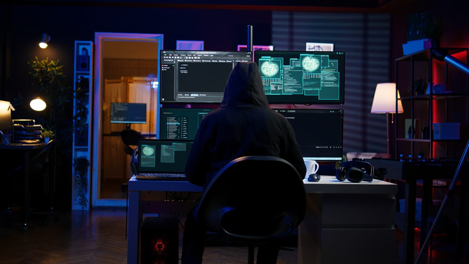 Hooded figure at a multi-monitor computer workstation, viewing code and technical graphics in a dark blue-lit room; tense, mysterious atmosphere.