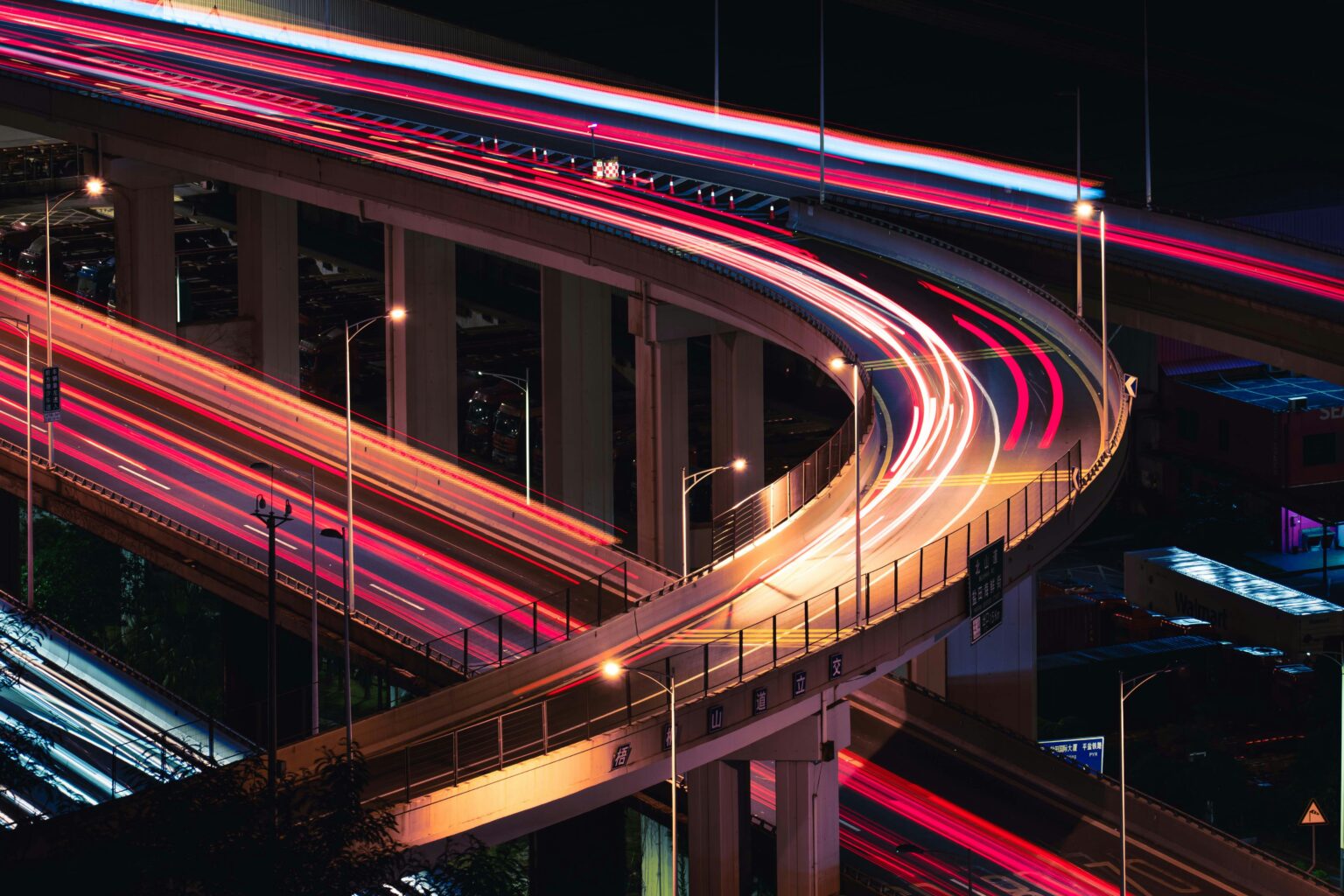 Nighttime highway interchange curves through a city, captured in long exposure with vivid red and white light trails and glowing streetlights