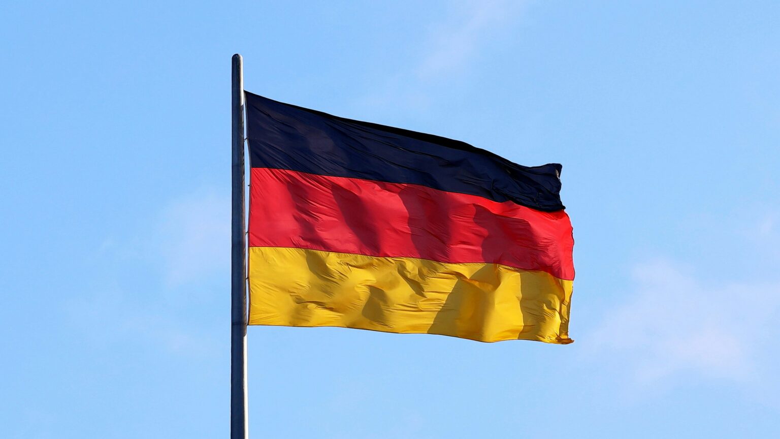 German flag with black, red, and gold horizontal stripes waves on a pole against a bright blue sky, conveying national pride and freedom
