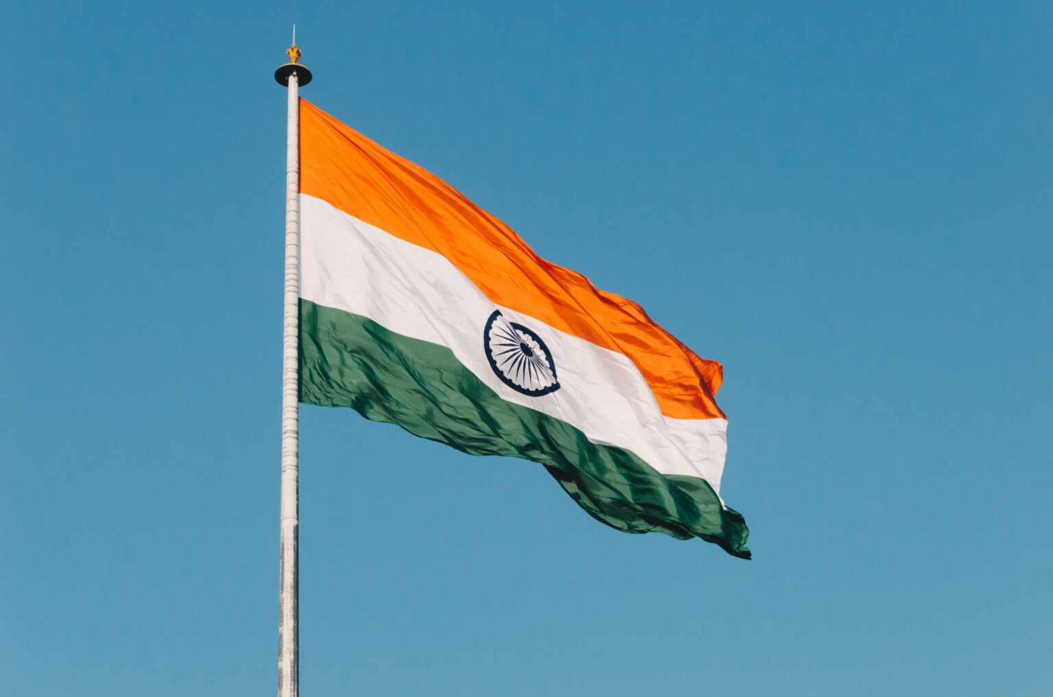 India’s saffron, white, and green tricolor flag, with the navy Ashoka Chakra, waves on a tall pole against a clear blue sky in bright sunlight, conveying national pride