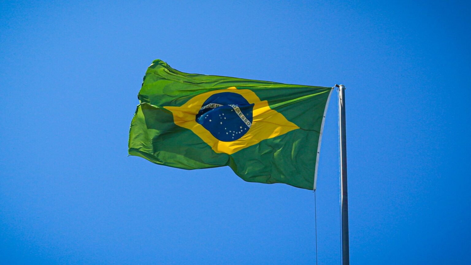 Brazilian flag waving on a tall flagpole against a clear blue sky, conveying national pride, freedom, and a bright, uplifting mood