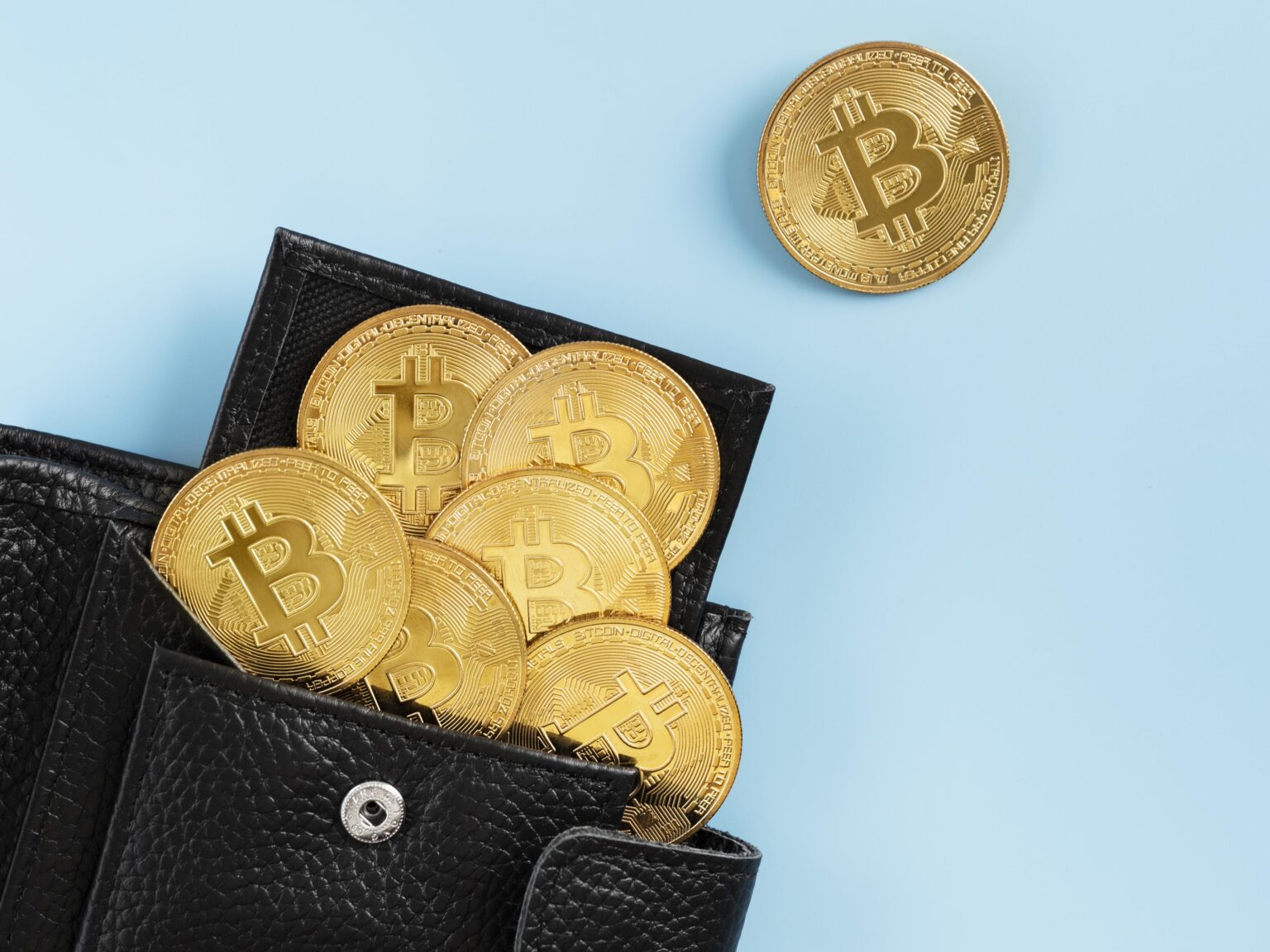 Black leather wallet filled with gold Bitcoin coins, with one Bitcoin coin above it on a blue background